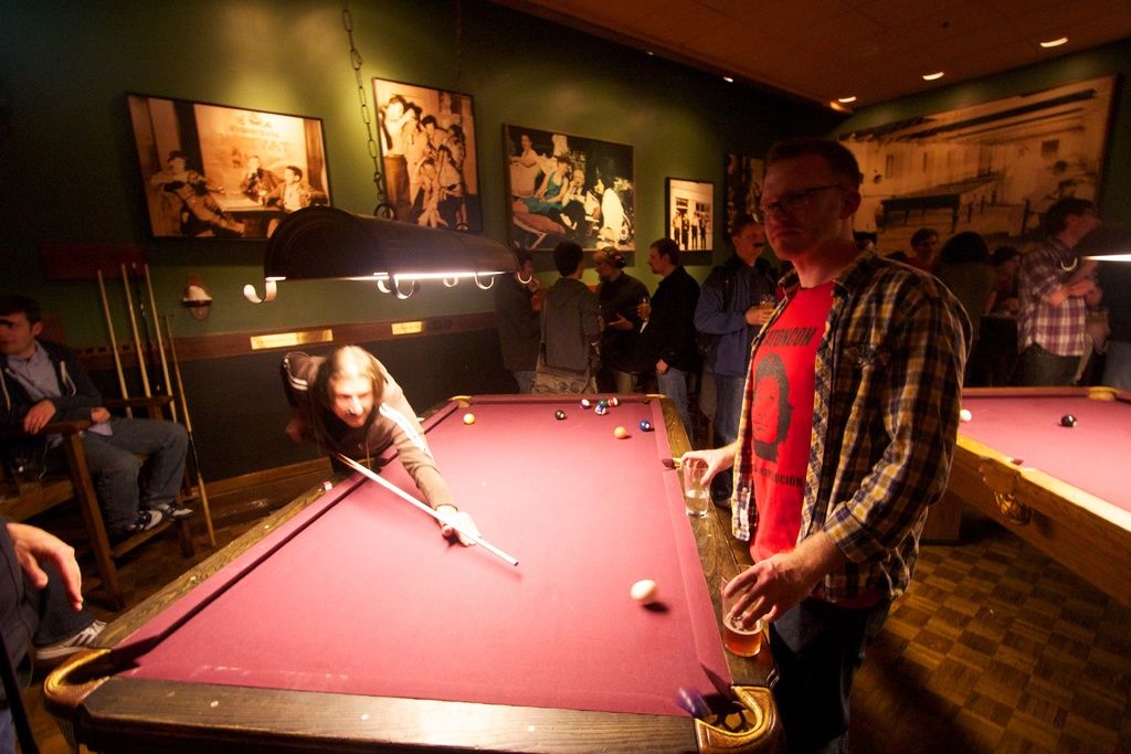 The picture is inside a club, there are two snooker tables, on the first table two people are...